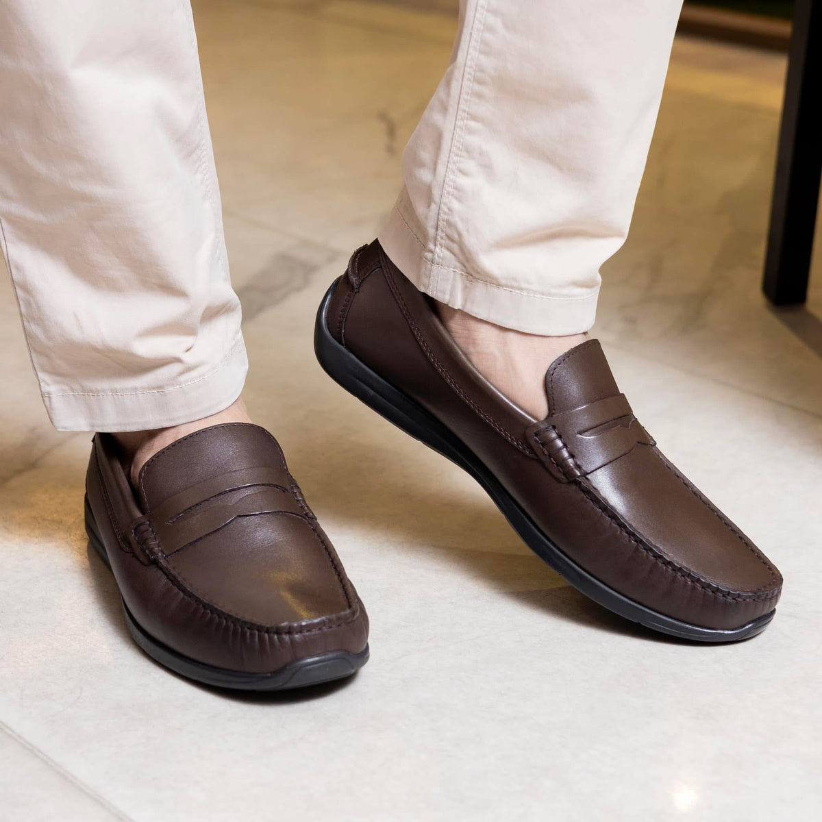 A man wearing brown leather dress loafers with a cushioned inner sole, standing on a light-colored floor.